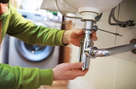 A plumber fixes pipes under a sink, likely a repair to remedy a home plumbing emergency.