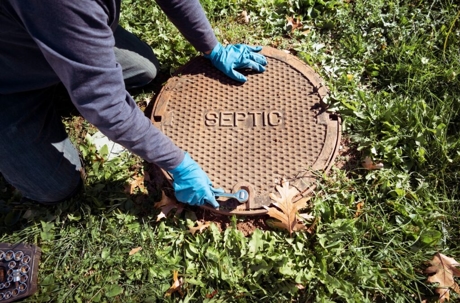 A technician uses a tool to open a septic tank lid in the ground. The metal lid is labeled "SEPTIC".