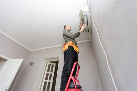 A technician is standing on a ladder while installing an air conditioning unit on a wall.