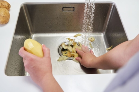 A person is peeling potatoes into a sink with a garbage disposal