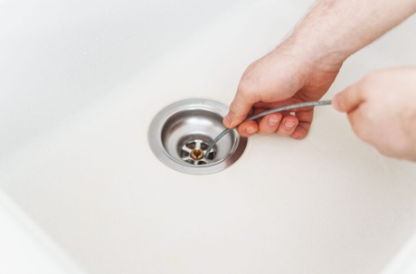 A person is using a plumbing snake to clear a clogged drain in a white sink.