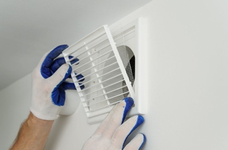 Hands in blue gloves install a white vent cover on a white wall.
