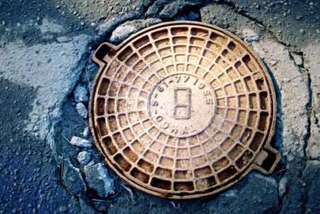 A worn metal manhole cover with a grid pattern sits in cracked pavement, likely related to sewer line inspections