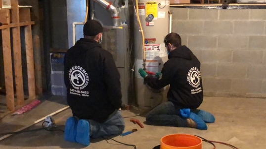 Two plumbers kneel and service a residential water heater, likely flushing the tank