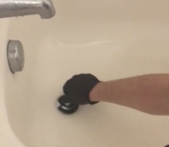 A gloved hand is seen removing a black drain cover from a bathtub