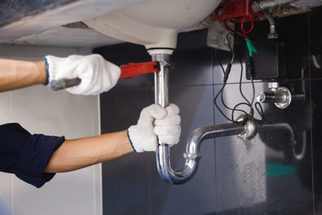 A plumber wearing gloves tightens a pipe under a sink with a wrench to repair it.