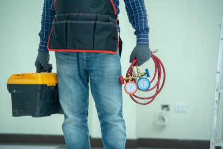 A tradesman in work clothes holds a toolbox and pressure gauges.