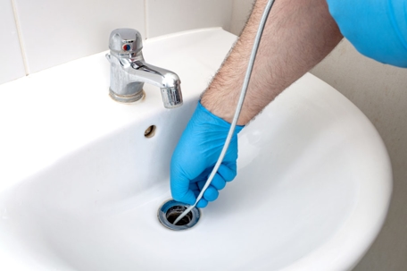 A plumber wearing blue gloves is clearing a clogged bathroom sink drain using a plumbing snake tool