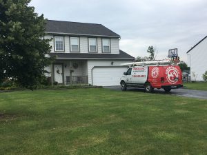 A two-story home with a white exterior and black roof. Parked nearby is a red and white Emergency Heating Services van