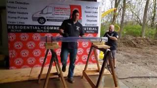 Two plumbers stand behind a long pipe supported by wood sawhorses.