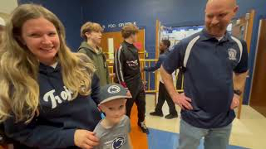 A still frame from a video features a group of people in a hallway, possibly a school, including a child in a Penn State hat, and others dressed in team apparel.