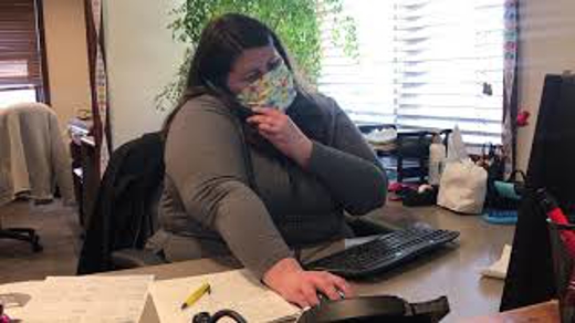 A woman sits at an office desk wearing a mask, speaking on the phone.