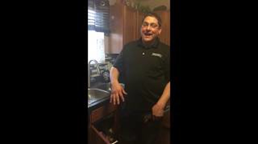 A man in a black shirt stands by a kitchen sink, possibly demonstrating plumbing work.