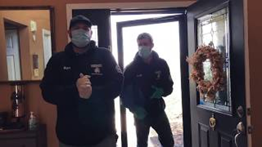 Two men stand at a front door, wearing masks and gloves.