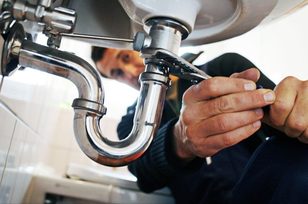 A plumber, under a sink, is fixing a pipe with a wrench.