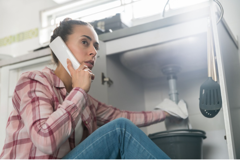 The Most Common Plumbing Problems Every Homeowner Should Know 4 Woman is on the phone with a plumber while inspecting a leaky pipe under her sink
